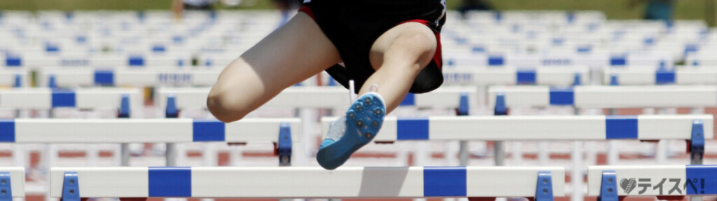陸上競技場のトラックで、並んでいる白と青のハードルを勢いよく飛び越えている選手の脚部のアップ