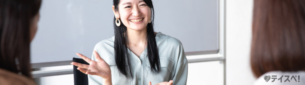 明るい室内で、水色のブラウスを着て大ぶりのイヤリングをつけた女性が、笑顔で相手に向かって手を広げながら話しています。