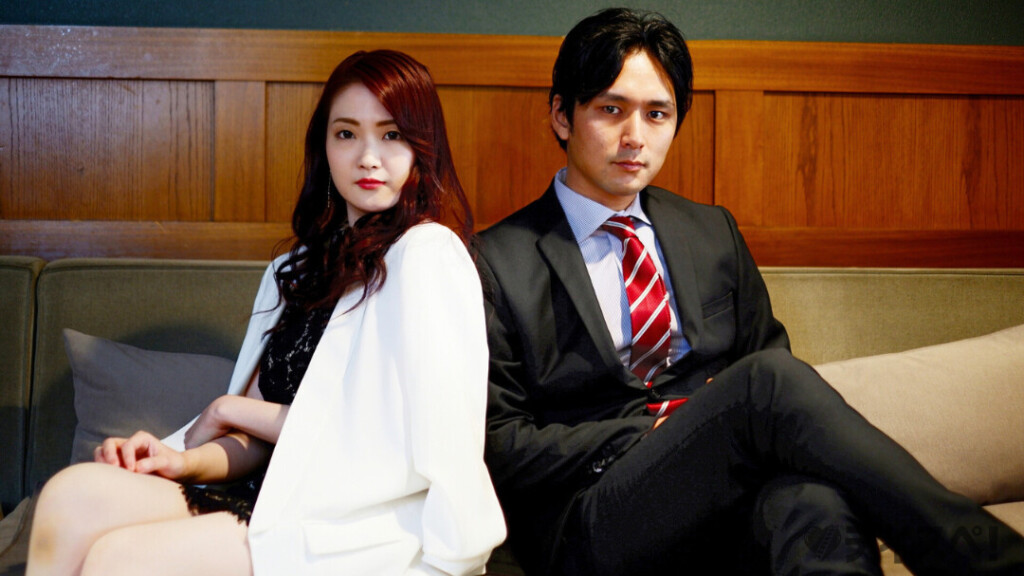 木目調の壁を背景に、白いジャケットを着たクールな表情の女性と、スーツに赤いネクタイを締めた男性がソファに背中合わせで座っています。