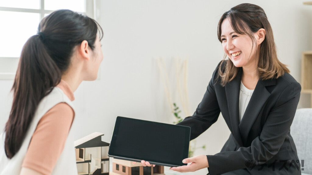 明るい室内で、黒いスーツ姿の女性が笑顔でタブレットの画面を相談者に見せています。テーブルの上には住宅の模型が置かれ、親身にアドバイスを行っているシーンです。