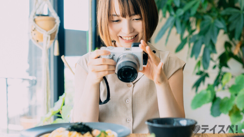 明るい室内で、ベージュの服を着たボブヘアの女性が笑顔で白いカメラを構え、テーブルの上の料理を撮影しようとしている様子。