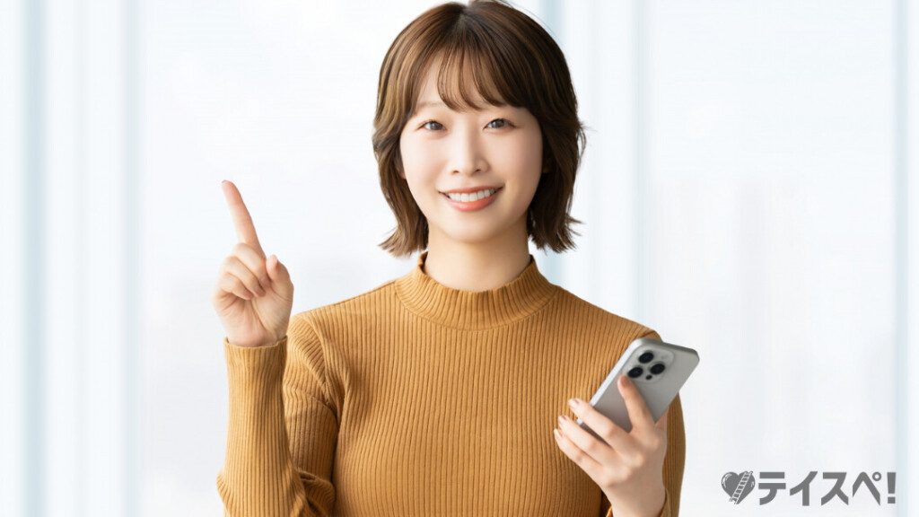 茶色のニットを着た若い女性が、左手にスマートフォンを持ちながら、右手の指を一本立てて「ポイント」を示すようににこやかに微笑んでいます。